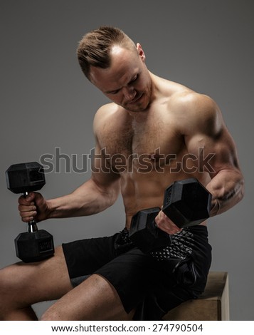 Muscular bodybuilder guy sitting on chair and doing exercises with dumbell on grey background