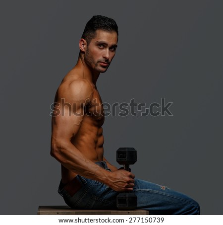 Shirtless guy sitting on a podium and holding one dumbell. Isolated on grey background