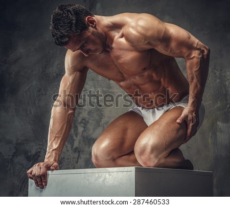 Bodybuilder guy posing on white podium on his knees. Isolated on grey background.