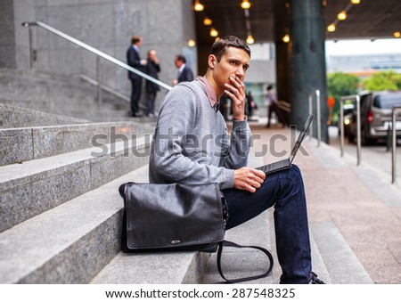 Smart guy in grey shirt and jeans sitting on stairs and working with laptop