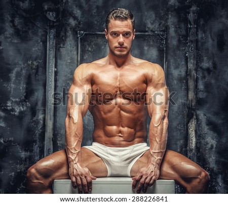 Awesome shirtles muscular guy sitting on white podium. Isolated on grey background.