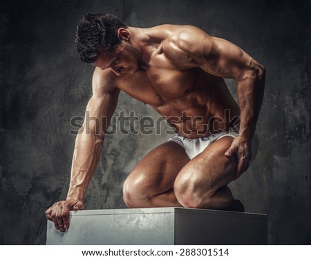 Muscular guy in white panties standing on his knees on white podium in studio.