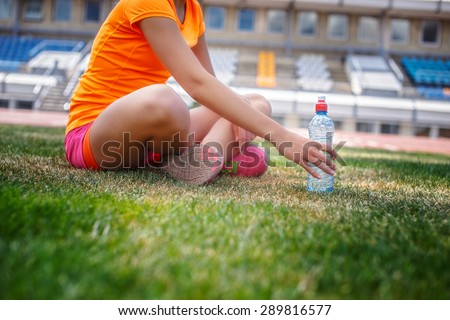 Girl in sportswear with bottle of water sitting on stadium grass.