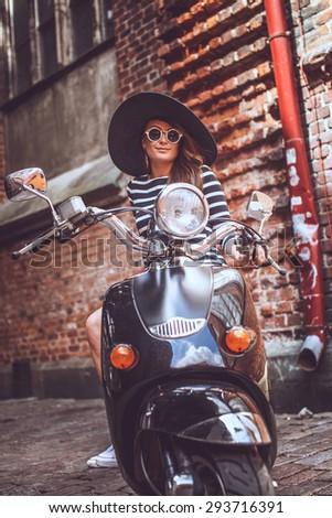 Female in black and white dress and summer hat sitting on her  scooter on red bricks wall background.