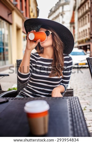 Cute female in big summer hat and sunglasses drinking coffee in summer cafe.