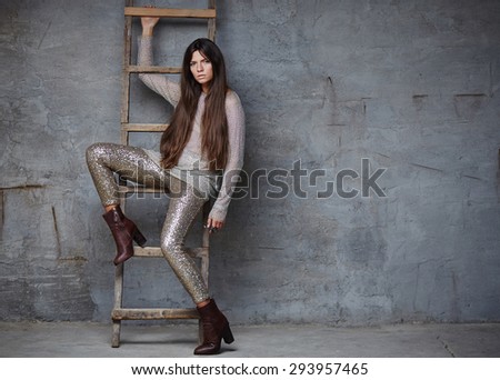 Slim female with long brown hair in shiny clothes posing near wood ladder.