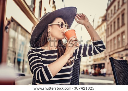 Beautiful young lady in big summer hat and in dress with stripes drinking coffe outside in summer cafe