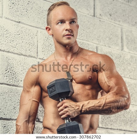 Shirtless muscular guy with dumbells posing near grey wall wfrom bricks.