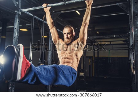 Shirtless man with deard in blue pants doing exersices on horizontal bar in a gym.