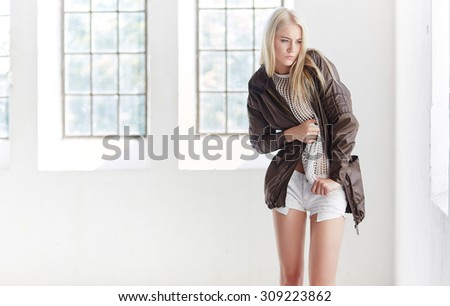 Full body portrait of sexy blond female in white dress and black jacket over window and white wall.