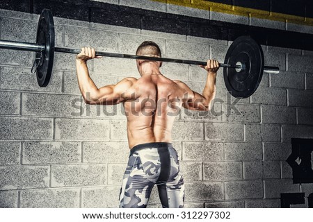 Shirtless man in military pants doing exercises with barbell over grey background. View from back.