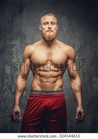 Shirtless muscular bearded man in red pants posing in studio over grey background.