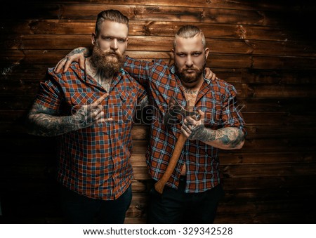 Two mans with beards and tattoo posing over wooden wall. One of them holding axe.