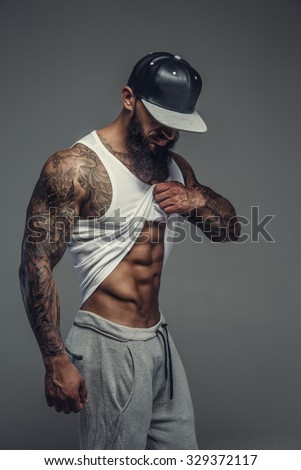 Muscular tattooed man with beard looking down and posing on camera. Isolated on grey background.