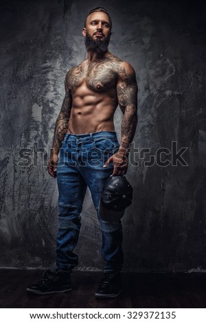 Full body portrait of muscular tattooed bearded man in blue jeans over grey wall.