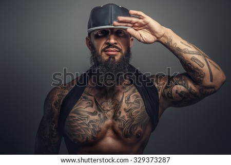 Brutal tattooed man in black cap posing in studio. Isolated on grey background.