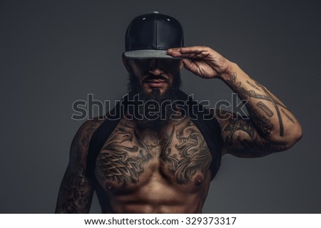Brutal tattooed man in black cap posing in studio. Isolated on grey background.