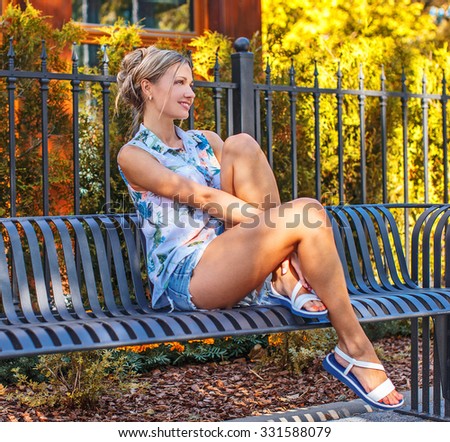 Sexy female in jeans shorts sitting on the bench with green bushes and houses on background.