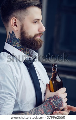 Bearded man with tattooes on his arm in a white shirt drinking beer.