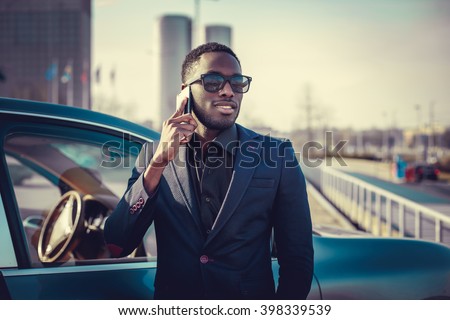 Casual black man in a suit talking by smartphone near a car.