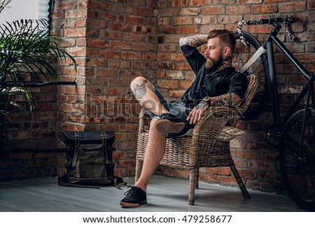 Portrait of a bearded hipster male relaxing in a chair with fix bicycle and the wall from the red brick background.