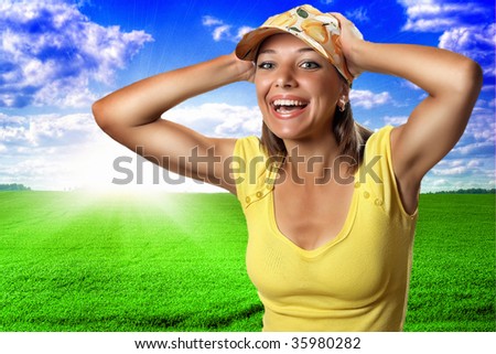 Young happy woman in yellow shirt and yellow peak cap with hands near head opposite abstract field and sky