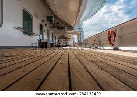 Very low ground view of a long cruise ship deck, with white life boat above and life saver ring on right side, with a dramatic blue, cloudy sky.