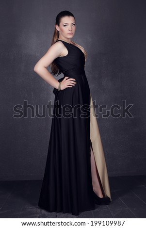 Young brunette lady in black ang golden dress posing on grey background. Fashion studio shot of beautiful woman with makeup and hairstyle wearing evening dress