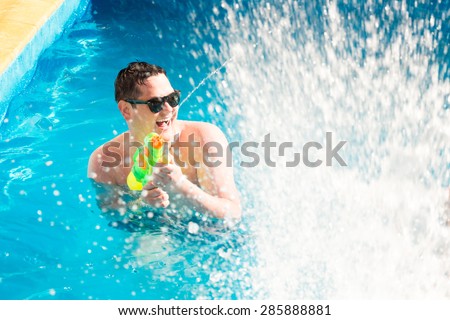 Joyful man with water gun in blue clear swimming pool with splashes.