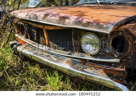 One headlight-Photograph of an old decaying car in a junk yard, that has lots of rust and one remaining headlight.
