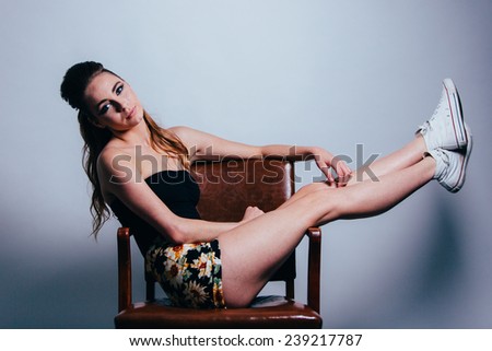 Studio Portrait of Young Attractive Woman sitting on brown leather chair sideways with legs sticking straight up head tilted
