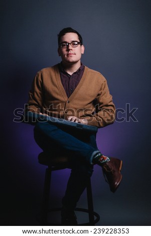 portrait of a young man in sweater vest sitting in chair holding a keyboard leg over the other