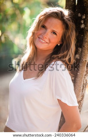 Beautiful Blonde Young Woman leaning on tree zoom vertical slight smile