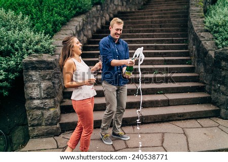 cute couple exploding a champagne bottle in front of stone steps laughing variation
