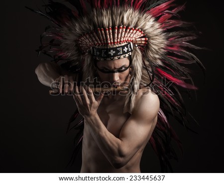 Portrait of the naked indian strong man posing with traditional native american make up with flute and headdress looking at the camera. Close up desaturated studio shot