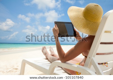 Woman holding tablet computer sitting on the beach in deck chair and taking sun bath