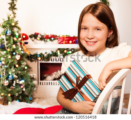 Happy girl sitting by fireplace and Christmas tree with present