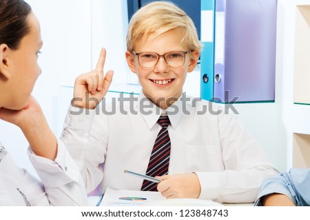 Kid playing adult with his friends sitting in the office pointing his finger up and smile as he have an idea