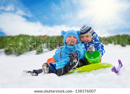 Two little boys brothers sitting on the snow and playing together on winter day