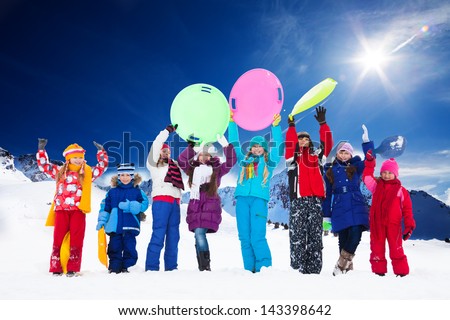 Row of large group of kids, friends, boys and girls standing together outside in snow, waiving hands, many holding sled