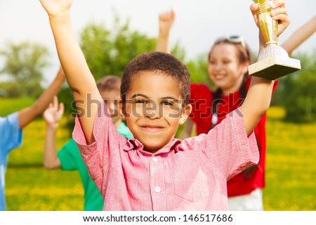 Close shoot of black happy smiling little boy holding prize cup with other kids cheering on the back