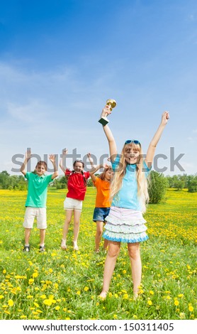 Portrait of happy little girl holding prize in lifted hands cup with group of kids on background