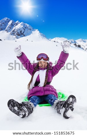 Happy girl sliding on sled with her hands lifted, wearing ski mask, with mountains on background