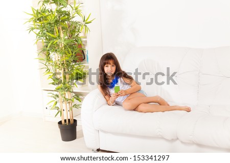 Happy shy smiling little girl with cell phone SMS her friends laying on the white leather couch