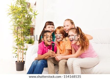 Group of five happy diversity kids, boys and girls, singing together sitting on the coach in living room