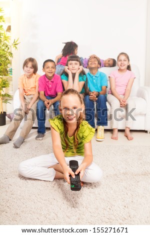 Little girl with TV remote control sitting on the floor in living room with her friends sitting on the coach on background