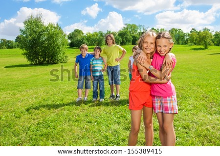 Two happy nine years old girls standing in the park hugging with group of guys friends standing on the background in the park on sunny day
