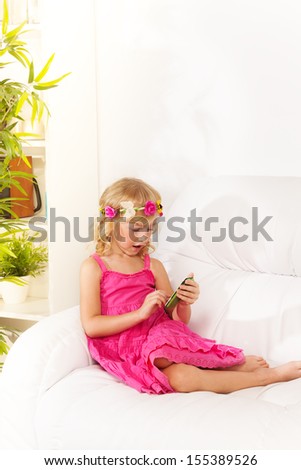 Little girl wearing wreath text her friends on social network sitting on the coach in living room