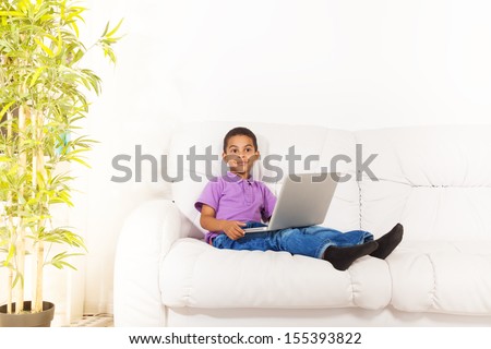 Smiling black little boy sitting on the white coach with laptop at home