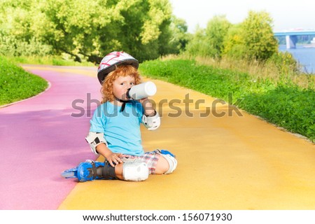 Happy thee years old boy sit with rollerblading and drink water from bottle in the park on sunny summer day
